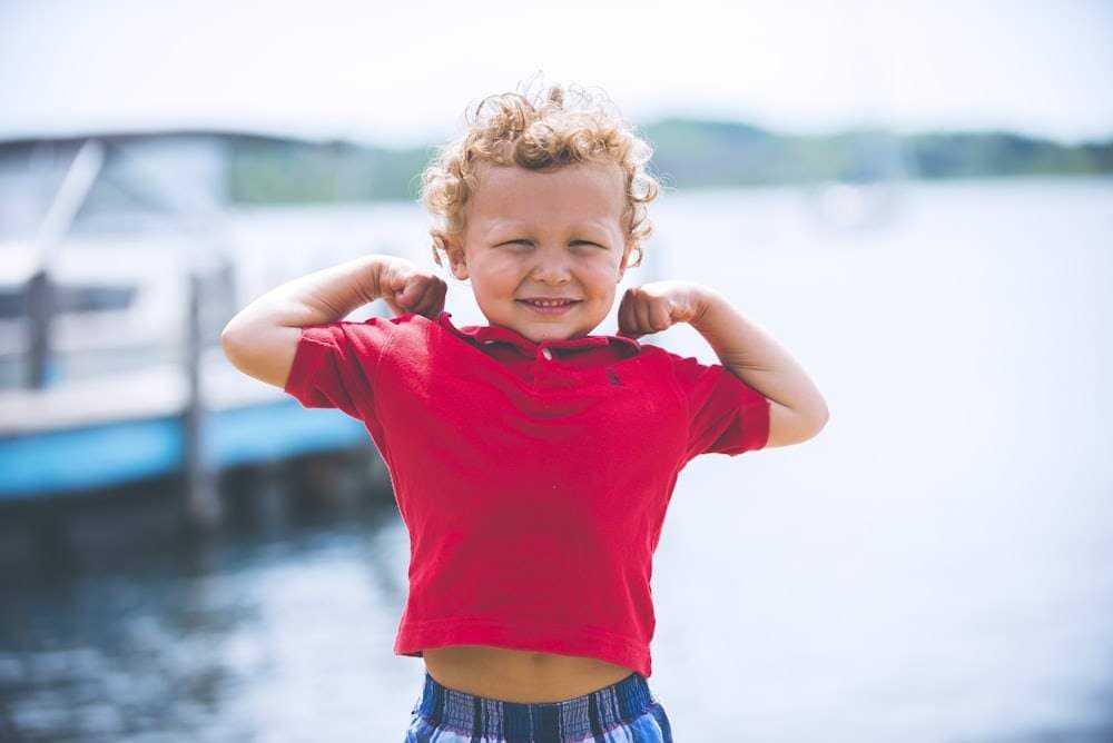 A toddler outdoors flexing both arms and grinning